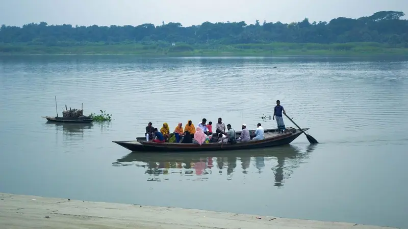 শান্ত নদীতে মাঝির বৈঠা বাওয়া যাত্রী বোঝাই একটি কাঠের নৌকা, দূরে সবুজ গাছের সারি