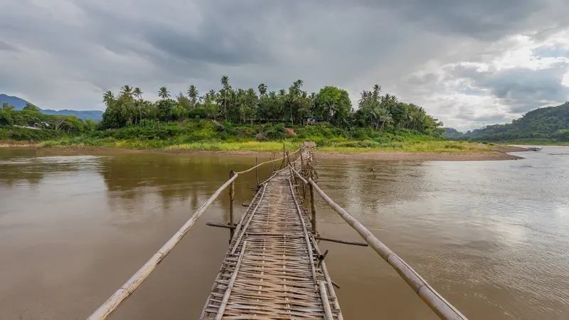 ঘোলা নদীর ওপর দিয়ে ওপারের সবুজ গাছপালার দিকে চলে যাওয়া একটি লম্বা কাঠের সাঁকো