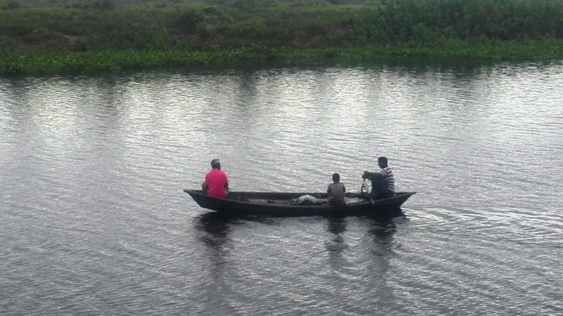 খালের শান্ত পানিতে তিনজন মানুষ নিয়ে ভেসে চলা একটি ছোট নৌকা