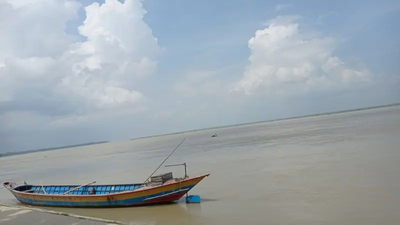 নদীর তীরে ভিড়ে থাকা একটি রঙিন কাঠের নৌকা, সামনে বিস্তীর্ণ পানি আর মেঘলা আকাশ